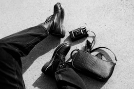 Black leather boots and dark jeans are prominent on a textured ground. A vintage camera and a small, stylish black leather bag are placed nearby.