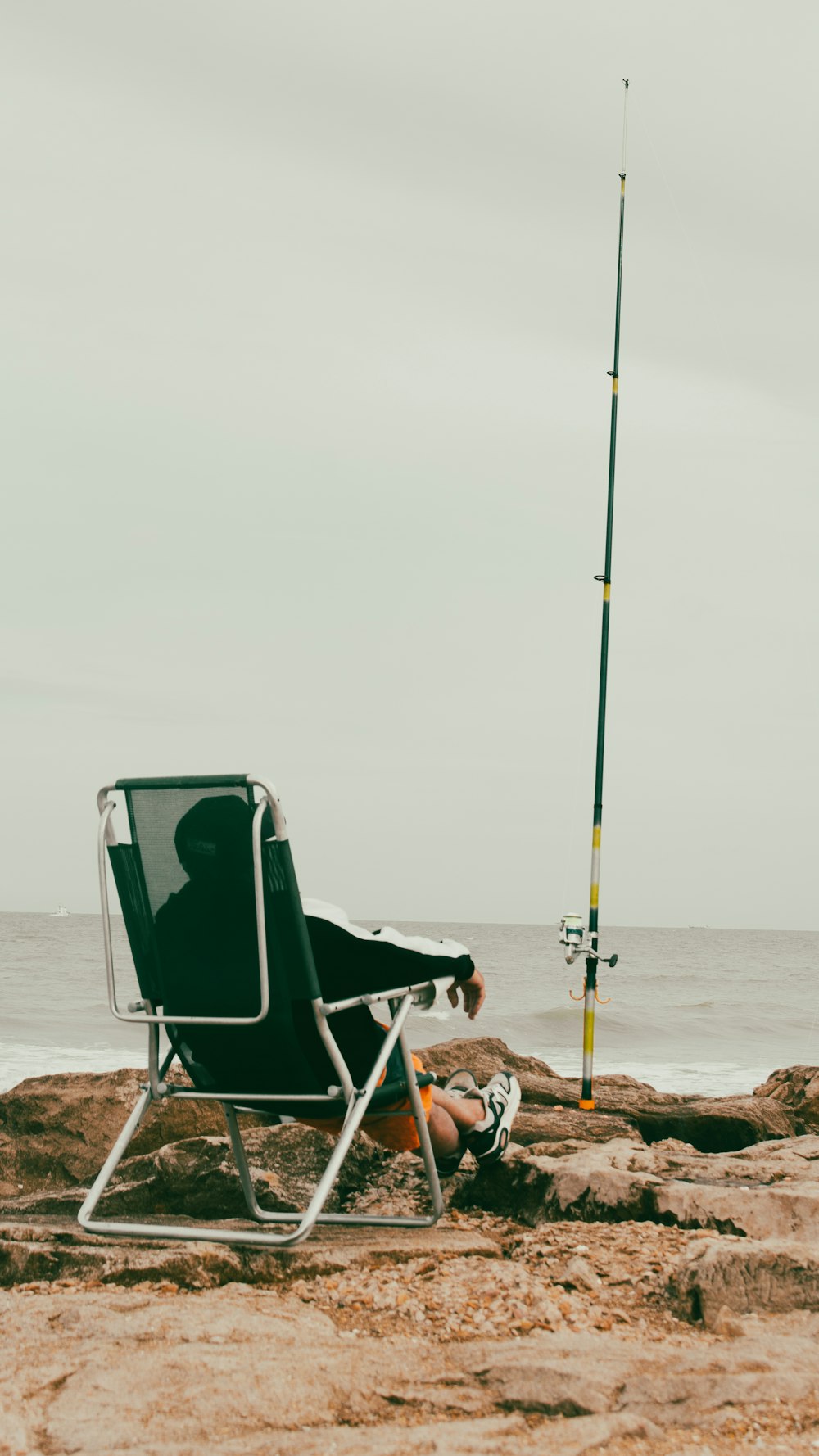 um homem sentado em uma cadeira na praia ao lado de uma vara de pesca