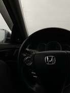 An interior view of a car, focusing on the steering wheel with a prominent Honda logo. The dashboard and part of the driver's side window are visible, with raindrops on the glass indicating rainy weather outside.