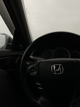 An interior view of a car, focusing on the steering wheel with a prominent Honda logo. The dashboard and part of the driver's side window are visible, with raindrops on the glass indicating rainy weather outside.