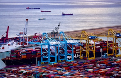 A bustling port filled with a multitude of colorful shipping containers stacked in rows. Several cranes in shades of blue and yellow are positioned above the containers, indicating active loading and unloading operations. Multiple ships are docked at the harbor, with more vessels visible in the calm water beyond the port. The scene conveys a sense of industrial activity and maritime trade.