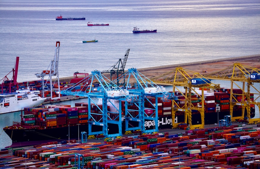 A bustling port filled with a multitude of colorful shipping containers stacked in rows. Several cranes in shades of blue and yellow are positioned above the containers, indicating active loading and unloading operations. Multiple ships are docked at the harbor, with more vessels visible in the calm water beyond the port. The scene conveys a sense of industrial activity and maritime trade.