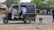 A parked auto rickshaw is situated on a dirt road with a hand-drawn heart symbol on its rear. Two people are seated inside, while a brown dog walks nearby. In the background, several people are passing by on foot and bicycles near small shops and a large tree.