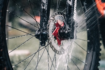 Close-up of a bicycle wheel being cleaned with water that splashes dramatically against the spokes and disc brake. The focus is on the metal rotor and red hub amidst the spray.