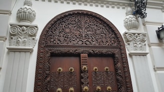 An intricately carved wooden door with an arch surrounded by decorative stone pillars, featuring floral and geometric designs. The wooden door is set into a white architectural facade, and there are ornate metal lanterns attached to the structure.