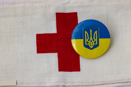 A red cross symbol is printed on a white fabric background. Next to it is a circular badge with the Ukrainian coat of arms in blue on a yellow background.