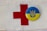 A red cross symbol is printed on a white fabric background. Next to it is a circular badge with the Ukrainian coat of arms in blue on a yellow background.