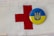 A red cross symbol is printed on a white fabric background. Next to it is a circular badge with the Ukrainian coat of arms in blue on a yellow background.