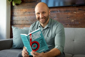 A man with a shaved head and beard is sitting on a gray sofa, smiling while holding a book titled 'Six Second Stories.' The setting has a cozy feel with a wooden wall behind and a plant visible in the corner.