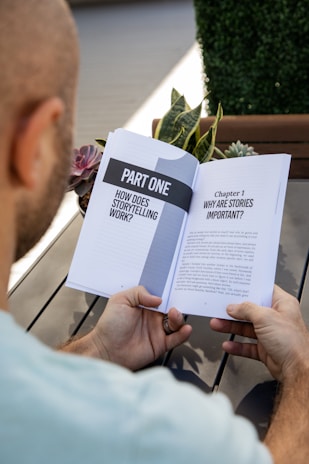 A person is holding an open book titled 'Part One: How Does Storytelling Work?' The book discusses the importance of stories. The setting includes a wooden table, plants, and a blurred outdoor background.