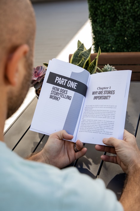 A person is holding an open book titled 'Part One: How Does Storytelling Work?' The book discusses the importance of stories. The setting includes a wooden table, plants, and a blurred outdoor background.