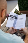 A person is holding an open book titled 'Part One: How Does Storytelling Work?' The book discusses the importance of stories. The setting includes a wooden table, plants, and a blurred outdoor background.