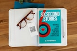 A wooden desk holds an open lined notebook with tortoiseshell eyeglasses and a QR code card resting on the pages. Adjacent to the notebook is a blue book titled 'Six Second Stories,' which focuses on maximizing impact with video storytelling. A silver pen is placed beside the notebook.