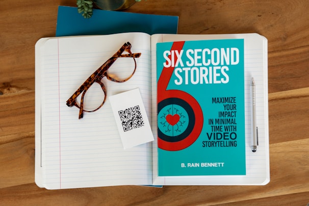 A wooden desk holds an open lined notebook with tortoiseshell eyeglasses and a QR code card resting on the pages. Adjacent to the notebook is a blue book titled 'Six Second Stories,' which focuses on maximizing impact with video storytelling. A silver pen is placed beside the notebook.
