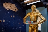 A gold humanoid robot is holding a tray with several stacked items. The wall in the background displays a dark blue space-themed mural with a large white spaceship and scattered star-like dots.