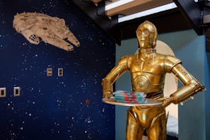 A gold humanoid robot is holding a tray with several stacked items. The wall in the background displays a dark blue space-themed mural with a large white spaceship and scattered star-like dots.