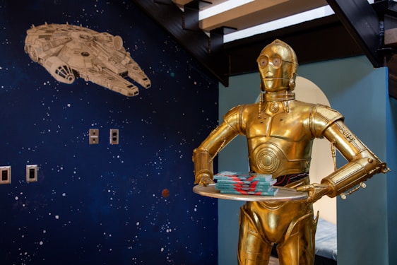 A gold humanoid robot is holding a tray with several stacked items. The wall in the background displays a dark blue space-themed mural with a large white spaceship and scattered star-like dots.