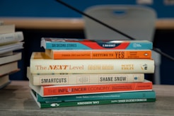 A stack of books is placed on a table with various titles visible, ranging from business negotiation and success strategies to creativity and philanthropy. The books are neatly arranged and the background includes another stack of books slightly out of focus.