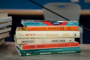 A stack of books is placed on a table with various titles visible, ranging from business negotiation and success strategies to creativity and philanthropy. The books are neatly arranged and the background includes another stack of books slightly out of focus.