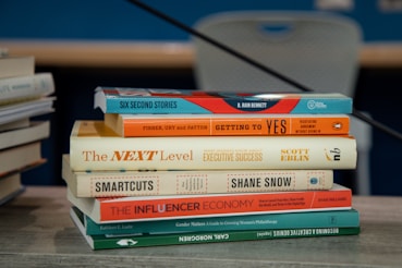 A stack of books is placed on a table with various titles visible, ranging from business negotiation and success strategies to creativity and philanthropy. The books are neatly arranged and the background includes another stack of books slightly out of focus.