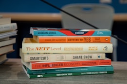 A stack of books is placed on a table with various titles visible, ranging from business negotiation and success strategies to creativity and philanthropy. The books are neatly arranged and the background includes another stack of books slightly out of focus.