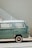 A vintage green Volkswagen camper van is parked beside a light-colored wall with a white metal staircase. The van's paint has a slightly worn appearance, and sunlight casts soft shadows on its surface.