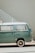 A vintage green Volkswagen camper van is parked beside a light-colored wall with a white metal staircase. The van's paint has a slightly worn appearance, and sunlight casts soft shadows on its surface.
