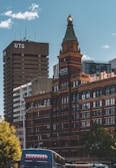 An urban scene featuring a red-brick historic building with a prominent clock tower topped with a green-capped spire. The background includes a modern high-rise building labeled 'UTS' and other contemporary structures. In the foreground, a bus with the label 'Warrigal' is visible, along with trees lining the street.