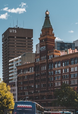 An urban scene featuring a red-brick historic building with a prominent clock tower topped with a green-capped spire. The background includes a modern high-rise building labeled 'UTS' and other contemporary structures. In the foreground, a bus with the label 'Warrigal' is visible, along with trees lining the street.