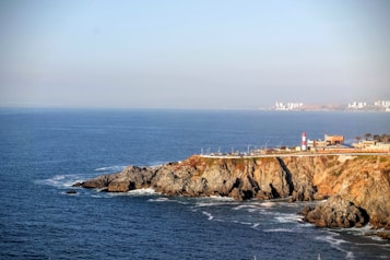 A scenic coastal landscape featuring rocky cliffs jutting into the sea, with gentle waves crashing against them. A lighthouse with red and white stripes stands prominently on the cliff. In the background, a cityscape with multiple tall buildings is visible across the water, under a clear blue sky.