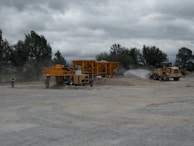 A construction site with heavy machinery, including a large yellow vehicle and equipment moving gravel. The area is dusty, with a worker in a safety vest walking nearby. Trees are visible in the background under an overcast sky.