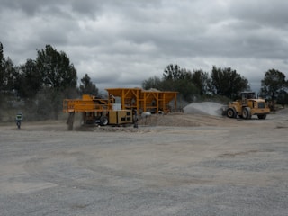 A construction site with heavy machinery, including a large yellow vehicle and equipment moving gravel. The area is dusty, with a worker in a safety vest walking nearby. Trees are visible in the background under an overcast sky.