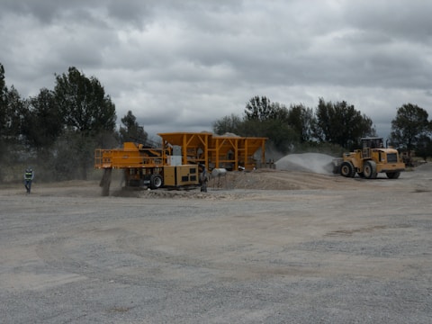 A construction site with heavy machinery, including a large yellow vehicle and equipment moving gravel. The area is dusty, with a worker in a safety vest walking nearby. Trees are visible in the background under an overcast sky.