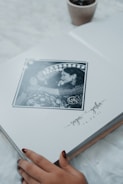 A photo album lies open to a page featuring a black and white photograph of a couple in an intimate pose, surrounded by an artistic border. Below the photograph are handwritten style names and a date. The corner of a small plant pot is visible in the upper part of the image, and a person's hand with painted nails is gently touching the bottom edge of the page.