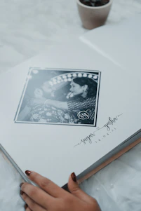 A photo album lies open to a page featuring a black and white photograph of a couple in an intimate pose, surrounded by an artistic border. Below the photograph are handwritten style names and a date. The corner of a small plant pot is visible in the upper part of the image, and a person's hand with painted nails is gently touching the bottom edge of the page.
