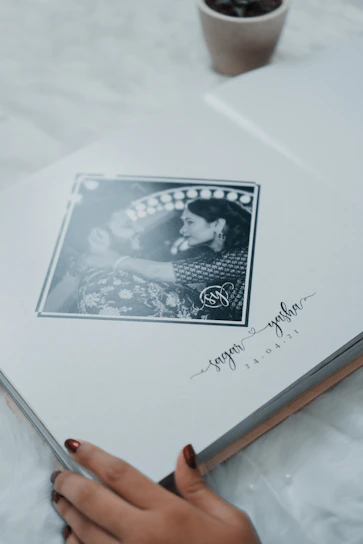 A photo album lies open to a page featuring a black and white photograph of a couple in an intimate pose, surrounded by an artistic border. Below the photograph are handwritten style names and a date. The corner of a small plant pot is visible in the upper part of the image, and a person's hand with painted nails is gently touching the bottom edge of the page.