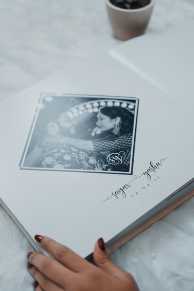 A photo album lies open to a page featuring a black and white photograph of a couple in an intimate pose, surrounded by an artistic border. Below the photograph are handwritten style names and a date. The corner of a small plant pot is visible in the upper part of the image, and a person's hand with painted nails is gently touching the bottom edge of the page.
