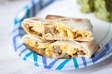 A close-up view of a breakfast burrito, cut in half, showcasing its interior filled with scrambled eggs, sausage, and melted cheese, positioned on a blue and white striped paper plate. A portion of green vegetables or garnish is visible in the background.