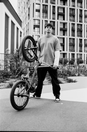A young person in casual clothing holds a BMX bike upright, standing against an urban backdrop featuring tall, modern buildings. The person is wearing a backward cap and has a confident posture. The scene is shot in black and white, adding a gritty, urban aesthetic.