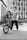 A young person in casual clothing holds a BMX bike upright, standing against an urban backdrop featuring tall, modern buildings. The person is wearing a backward cap and has a confident posture. The scene is shot in black and white, adding a gritty, urban aesthetic.