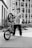 A young person in casual clothing holds a BMX bike upright, standing against an urban backdrop featuring tall, modern buildings. The person is wearing a backward cap and has a confident posture. The scene is shot in black and white, adding a gritty, urban aesthetic.