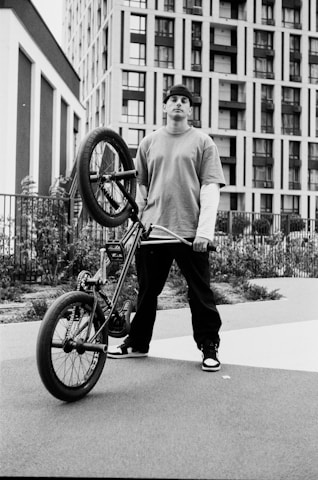 A young person in casual clothing holds a BMX bike upright, standing against an urban backdrop featuring tall, modern buildings. The person is wearing a backward cap and has a confident posture. The scene is shot in black and white, adding a gritty, urban aesthetic.