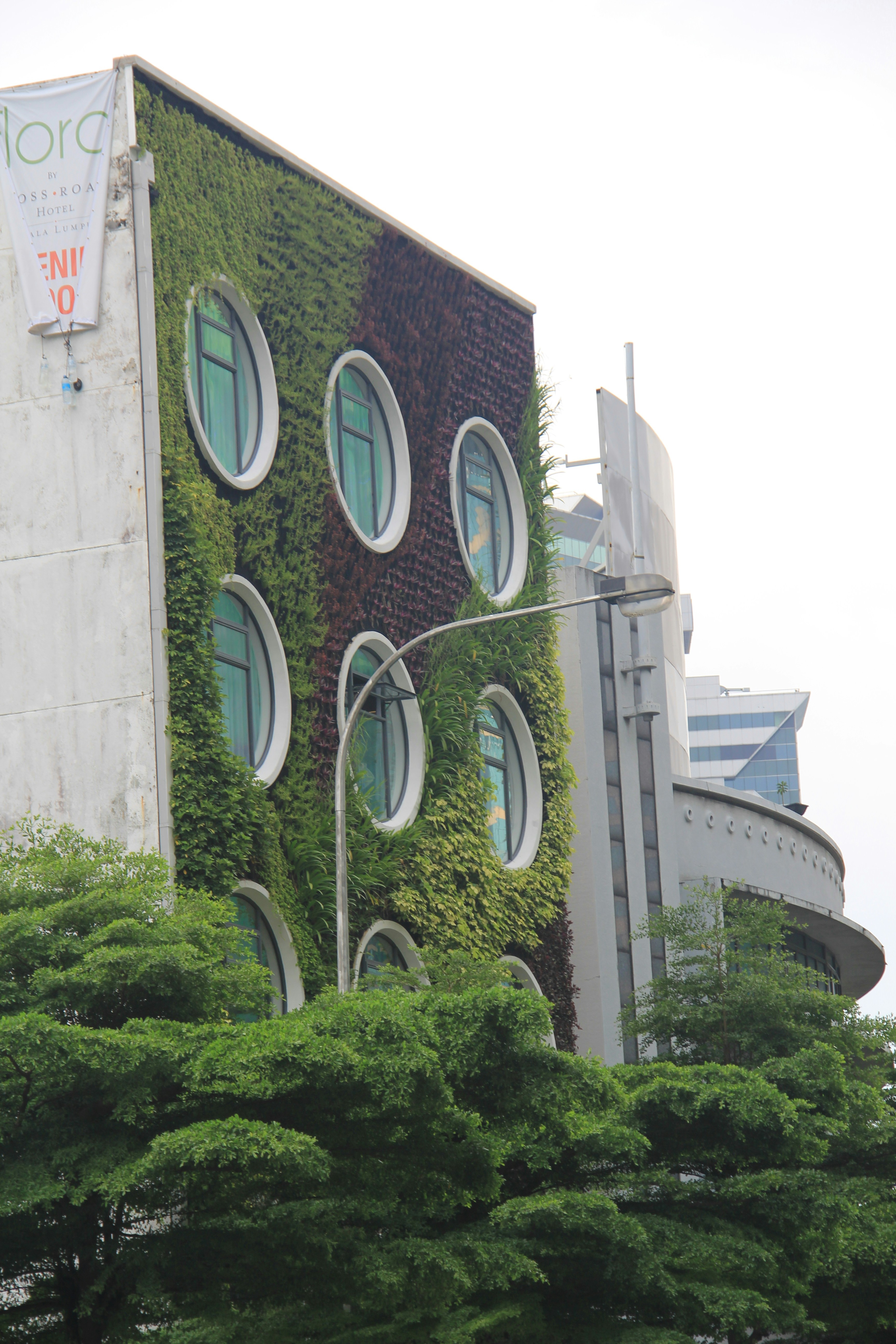 The building facade is covered with lush green vegetation, featuring round windows that resemble portholes. The structure appears modern and eco-friendly, with surrounding trees adding to the verdant atmosphere. Another building in the background complements the urban setting.
