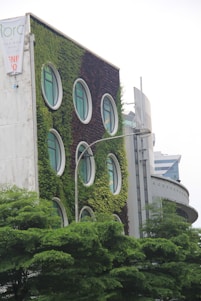 The building facade is covered with lush green vegetation, featuring round windows that resemble portholes. The structure appears modern and eco-friendly, with surrounding trees adding to the verdant atmosphere. Another building in the background complements the urban setting.