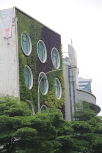 The building facade is covered with lush green vegetation, featuring round windows that resemble portholes. The structure appears modern and eco-friendly, with surrounding trees adding to the verdant atmosphere. Another building in the background complements the urban setting.