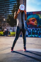A person with long braided hair is wearing a white cap and dark shiny pants, standing confidently on an urban street. Graffiti art is visible on a wall in the background, alongside a building with many windows.
