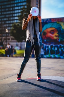 A person with long braided hair is wearing a white cap and dark shiny pants, standing confidently on an urban street. Graffiti art is visible on a wall in the background, alongside a building with many windows.