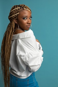 A person with long braided hair, wearing large hoop earrings, a white shirt, and denim jeans stands against a solid teal background. Their posture is turned slightly to the side, and they gaze intently towards the camera.