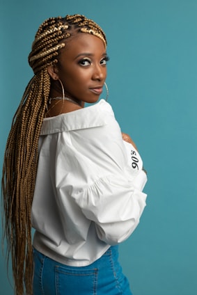 A person with long braided hair, wearing large hoop earrings, a white shirt, and denim jeans stands against a solid teal background. Their posture is turned slightly to the side, and they gaze intently towards the camera.
