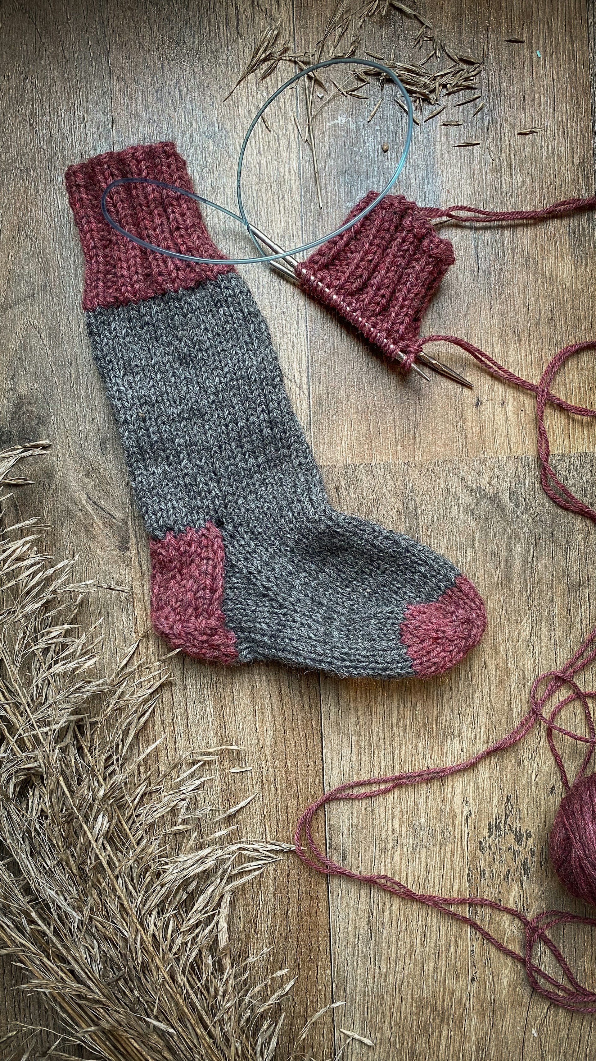 A handmade sock with a blend of gray and red yarns lies on a wooden surface next to a partially completed second sock still on the knitting needles. Dried grass or reeds are placed on the left side of the composition.